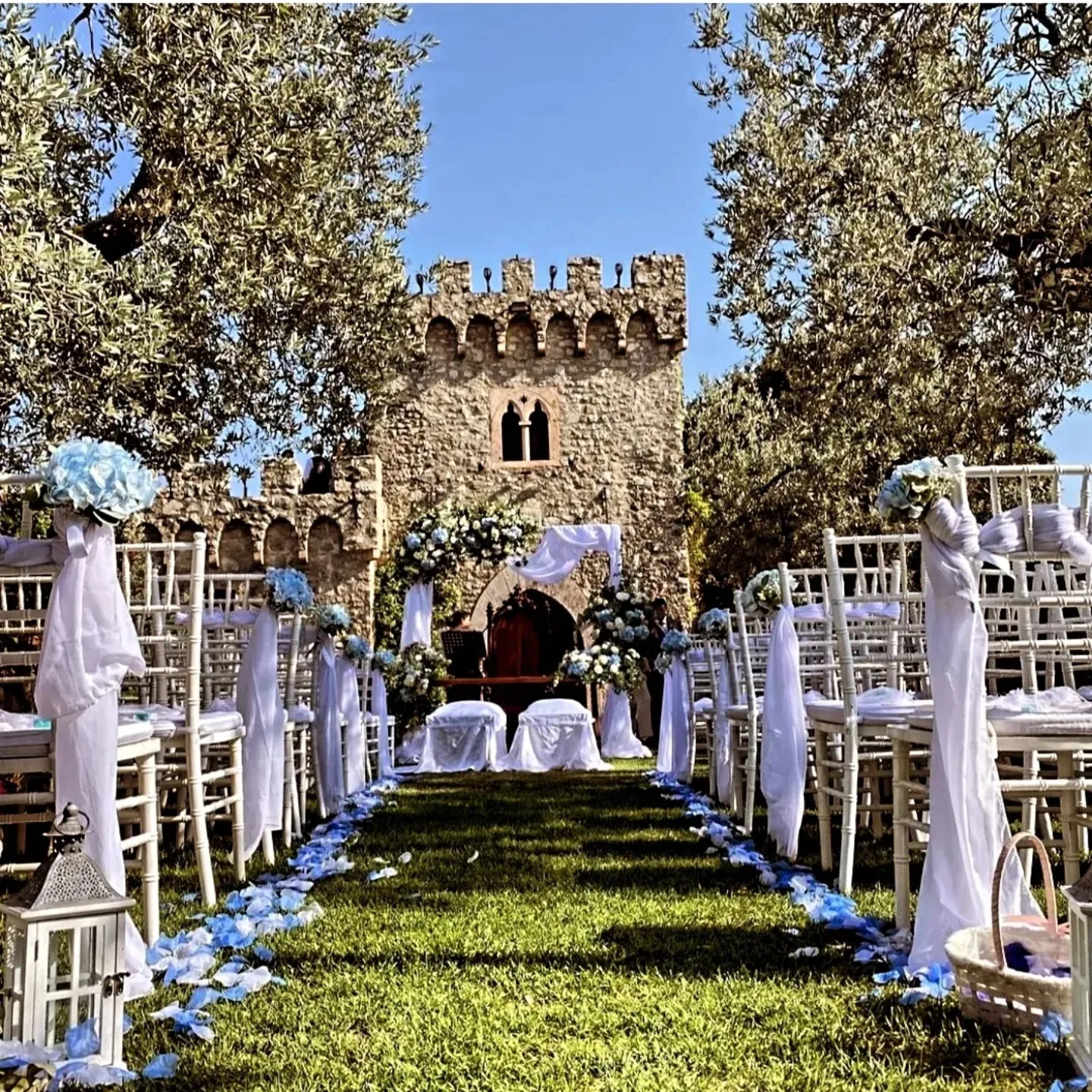 Cerimonia di matrimonio all'aperto davanti a un castello, con sedie bianche, fiori azzurri e petali sul prato